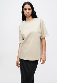 Bézs pamut póló laza szabással, rövid ujjú és kerek nyakkivágással. Kis fehér logó található a mellkasán, fekete szoknyával párosítva.