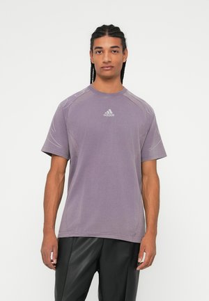Mees punutud juuste ja violetse Adidas T-särgiga, mustade pükste ja valge taustaga.