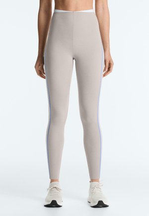 Slim fit bézs leggings halványkék oldalsávokkal, fehér tornacipőt viselő álló személyen, egyszerű háttér előtt.