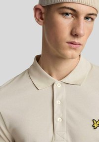Jonge man met een beige gebreide muts en een beige poloshirt met een gele adelaar logo, die iets opzij kijkt tegen een effen achtergrond.