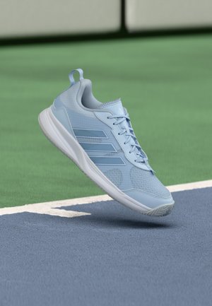 Pantof sport albastru deschis cu talpă albă plutind deasupra suprafeței unei teren de sport albastru și verde, în apropierea unei linii de delimitare albe.