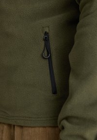 Close-up of olive green fleece jacket with vertical black zippered pocket and fabric loop pull on the chest.