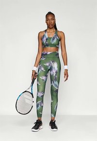 Mulher em roupa desportiva floral verde, segurando uma raquete de ténis, vestindo sapatilhas pretas e pulseiras brancas, em pé contra um fundo branco liso.