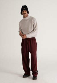 Gestickter, cremefarbener Rollkragenpullover mit gesprenkelter Textur; weinrote Jogginghose mit seitlichen Streifen; schwarze Mütze; schwarze Schuhe.