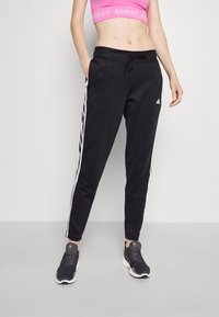 Schwarze Sporthose mit weißen Streifen an den Seiten, elastischem Bund mit Kordelzug, aus einem Polyester-Mix gefertigt, kombiniert mit schwarzen Turnschuhen.