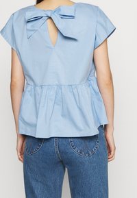 Top peplum azzurro chiaro con maniche a flusso, schiena a chiave di violino e dettaglio a fiocco. Il tessuto appare liscio con una finitura texturizzata. Abbinato a jeans blu.