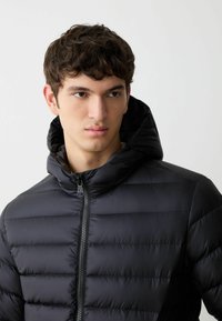 Giacca a vento nera con cuciture orizzontali, colletto alto e zip frontale. Il tessuto ha una texture liscia e lucida.