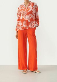 Rode bloemenblouse met witte patronen, lange mouwen en een v-hals. Gepaard met losse, feloranje broeken en platte sandalen.