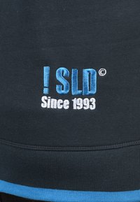 Zwarte sweatshirt met geborduurde tekst in lichtblauw en wit: "! SLD" en "Sinds 1993" met een blauwe accentstreep aan de zoom.