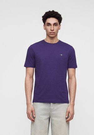 Jeune homme aux cheveux courts et bouclés, portant un t-shirt violet uni à manches courtes et un pantalon beige clair, debout devant un fond uni.