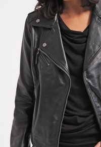 Femme portant une veste de motard en cuir noir sur un haut noir à col drapé, raccourci aux épaules et à la taille.