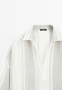 Chemise légère à manches longues avec des rayures verticales blanches et beiges, col ouvert et une petite étiquette de marque noire à l'intérieur du col.