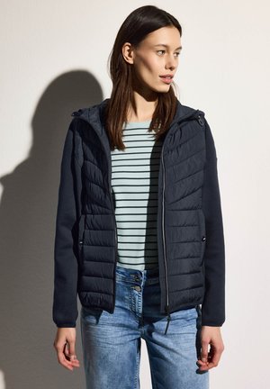 Navy puffer jacket with a hood, featuring quilted texture and fitted sleeves, layered over a light blue striped top and denim jeans.