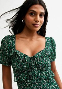 Robe florale verte avec des manches courtes bouffantes, un décolleté carré et des accents en volants. Conçue dans un tissu léger avec une taille cintrée.