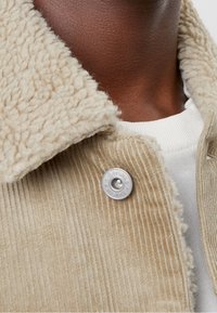Béžová kordurová bunda s huňatým ovčím límcem, stříbrnými knoflíky a strukturovanou tkaninou, pod kterou je vidět bílá košile.