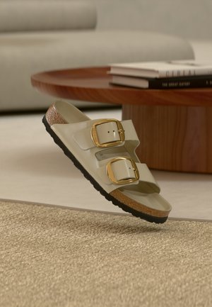 Light green sandal with two gold buckles, cork footbed, and a textured black rubber sole, set against a neutral background.