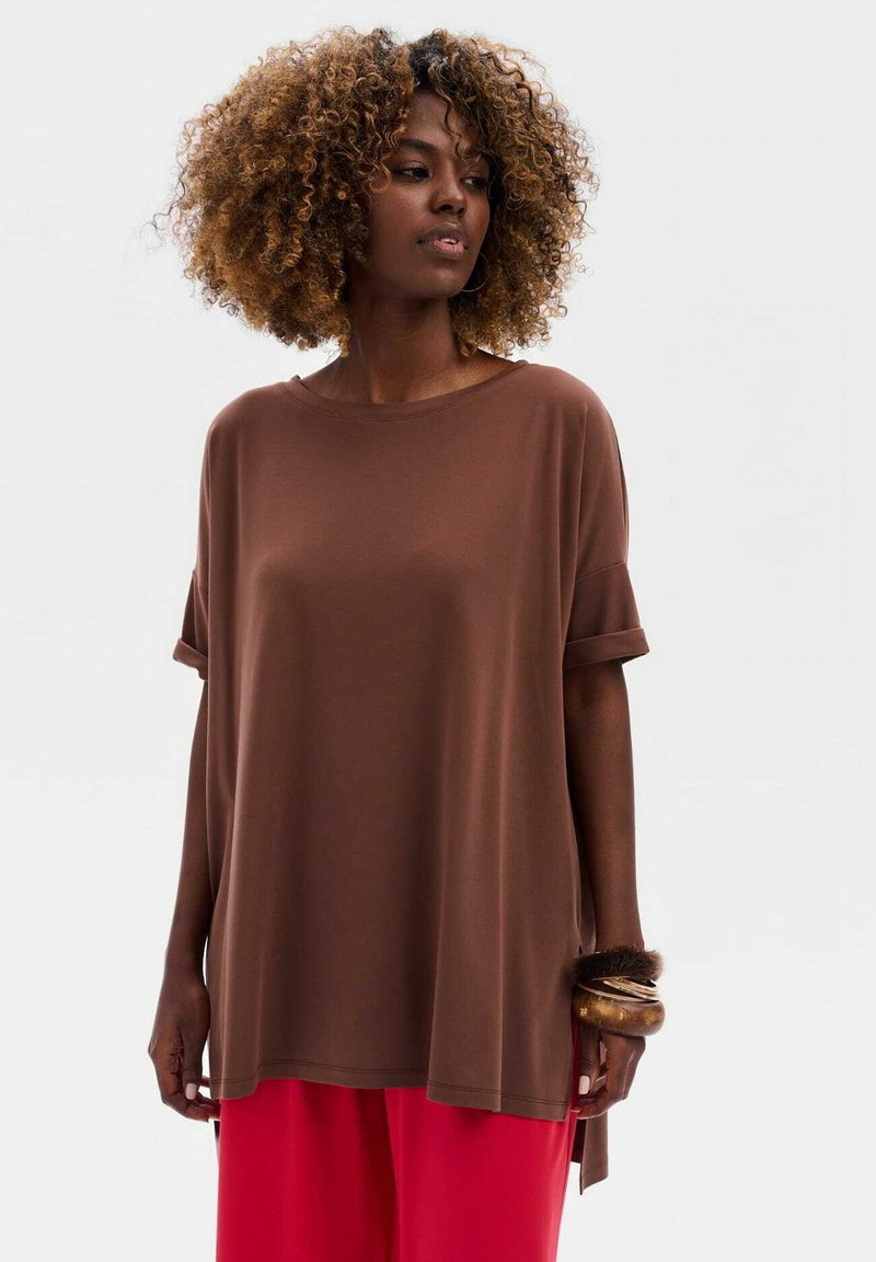 Brązowy oversize t-shirt z krótkimi, podwiniętymi rękawami, gładki materiał, w połączeniu z jasnoczerwonymi spodniami o szerokich nogawkach i teksturowaną bransoletką na nadgarstku.