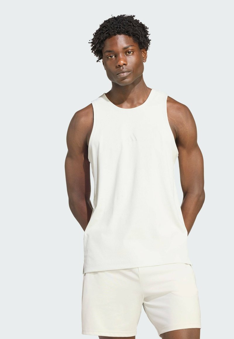 Athletic man with curly hair wearing a white sleeveless tank top and matching shorts, standing with hands behind his back.