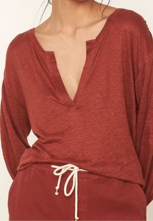 Femme portant un haut rouge ample à manches longues avec un col en V fendu et un pantalon rouge assorti noué avec un cordon blanc.