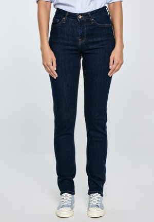 Persona, kas valkā tumši zilas pieguļošas džinsas, baltas un zilas kedas, un gaiši zilu pogājamu kreklu, stāv uz vienkrāsaina fona.