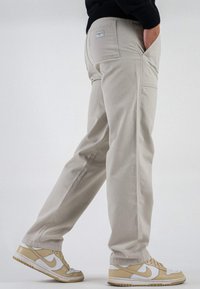 Pantaloni cargo color beige chiaro con tasche laterali, realizzati in cotone; abbinati a sneakers Nike beige e bianche, che mostrano un design e una texture semplici.
