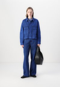 Blå denimjakke med to frontlommer, sammen med matchende vide bukser. Modellen holder en svart lærveske. Enkelt, uformelt design.