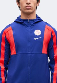 Άνδρας με μπλε και κόκκινο μπουφάν Nike Paris Saint-Germain με κουκούλα και μπροστινό φερμουάρ, εμφανιζόμενος από τους ώμους μέχρι το πηγούνι.