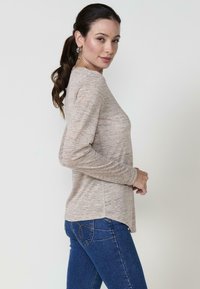 Langärmliges, hellbeiges Oberteil aus strukturiertem Strick mit abgerundetem Saum, kombiniert mit enganliegenden blauen Jeans, die dekorative Nähte auf den Gesäßtaschen aufweisen.