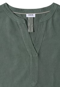 Chemise en lin verte avec un col rond et une ouverture en V fendue, avec les étiquettes "CECIL" et "LINEN" à l'intérieur du col.