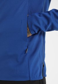 Giacca blu antivento con una consistenza morbida, tasca laterale con zip e una discreta etichetta sul bordo. Presenta maniche lunghe e un design aderente.