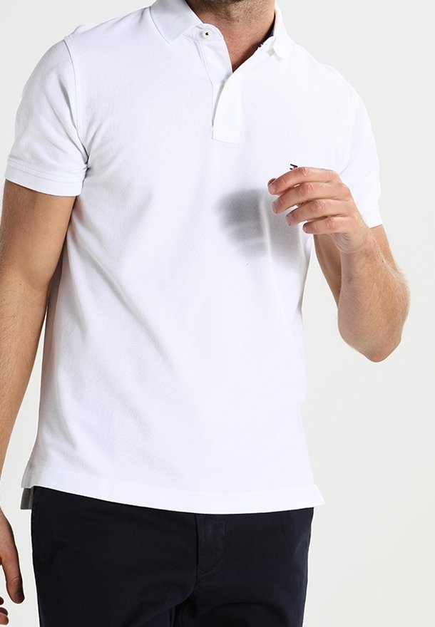 Homme portant un polo blanc uni à manches courtes et un pantalon foncé, une main levée près de la poitrine sur un fond clair.