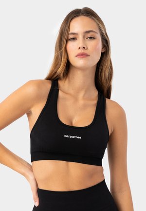 Mladá žena s dlhými vlasmi oblečená v čiernej športovej podprsenke Carpatree a zodpovedajúcich vysoko pásových legínach na jednoduchom pozadí.