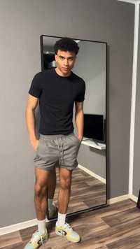 Egy fiatal férfi áll zsebre tett kézzel, fekete pólót, szürke rövidnadrágot, fehér zoknit és sárga tornacipőt visel egy nagy tükör előtt.