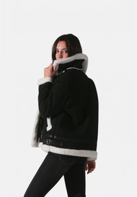 Veste en peau de mouton noire avec doublure en polaire blanche, col montant et accents en cuir. Comprend une fermeture éclair et des poches latérales.