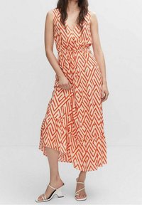 Femme portant une robe midi sans manches, à col en V, à motif géométrique orange et blanc, avec une taille nouée et des sandales à talons blancs à lanières.
