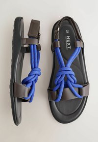Sandales plates noires avec des brides en corde bleue nouées et des accents en cuir marron, taille 6,5/40, étiquetées "NEXT Forever Comfort."