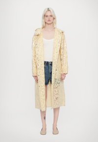 Trench-coat en dentelle jaune avec un motif floral, ouvert à l'avant, manches longues et épaulettes, porté sur un débardeur blanc et un short en jean.