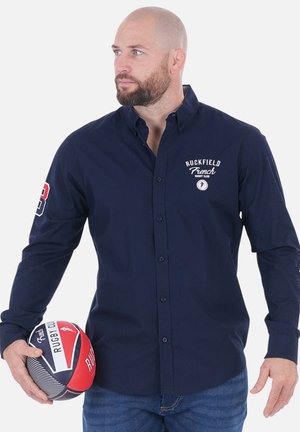 Chemise bleu marine à boutons avec le logo du Ruckfield French Rugby Club, manches longues, et design ajusté. Homme tenant un ballon de rugby rouge et bleu.