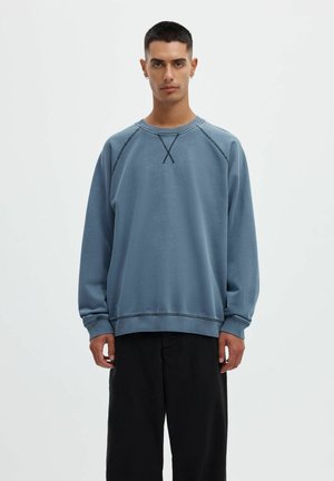 Jeune homme aux cheveux courts portant un sweat-shirt bleu délavé et un pantalon noir, debout sur un fond clair et uni.