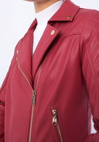 Červená kožená bunda z umělé kůže s prošívaným vzorem na ramenou, s kapsami na zip a zlatými detaily. Obsahuje klasický límec a hladký povrch.