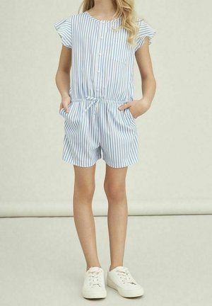 Enfant portant une combinaison courte à manches courtes rayée verticalement bleu et blanc avec des boutons à l'avant et des baskets blanches, debout avec les mains dans les poches.