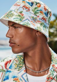 Chapeau seau avec un imprimé floral et océanique ; tissu blanc ; porté avec une chemise à fleurs ; chaîne en argent visible. Teint de peau foncé.