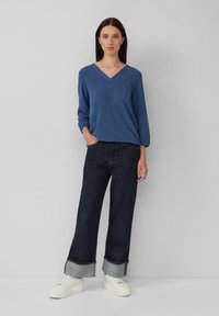 Femme se tenant contre un mur uni, portant un pull bleu à col en V, un jean large foncé avec des ourlets et des baskets plateformes blanches.