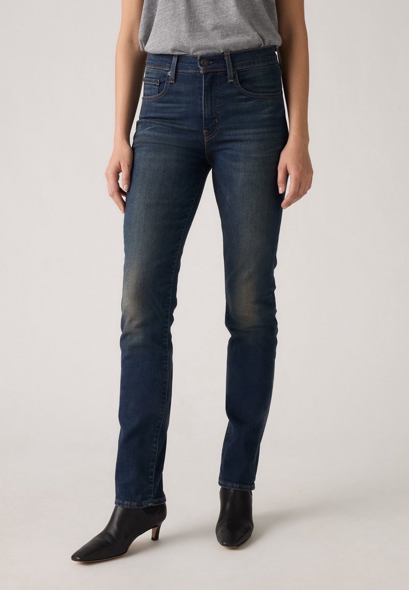 Femme portant un jean slim bleu foncé avec un léger effet délavé, un top gris et des bottines à talons pointues noires, debout sur un fond uni.