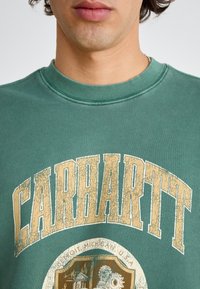 Zelená mikina s žebrovaným výstřihem, s velkým, popraskaným zlatým potiskem "CARHARTT" a kruhovou grafikou na spodní části.