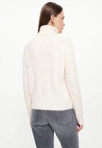 Crème kabelgebreide trui met een hoge kraag. Kenmerkt zich door een ribbeltextuur en een cropped pasvorm. Gecombineerd met hoge taille grijze jeans.