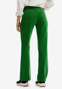 Jeans acampanados verdes de denim, con una textura suave, doble bolsillo trasero y una cintura alta, combinados con zapatillas negras y crema.