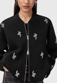 Sort zip-up jakke med ribbet tekstur, der har sølvsnegl broderi i forskellige positioner. Høj krave og sidelommer er synlige.