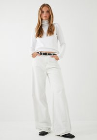 Witte coltrui gecombineerd met wijde witte jeans. Zwarte riem met zilveren gesp accent. Model staat met handen in de zakken.
