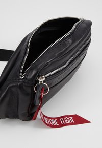 Musta nahkalaukku, jossa on tekstuuripinta, hopeinen vetoketju ja punainen nauhatagi, jossa lukee "POISTA ENNEN LENNON ALKUA." Sisus näkyvissä.